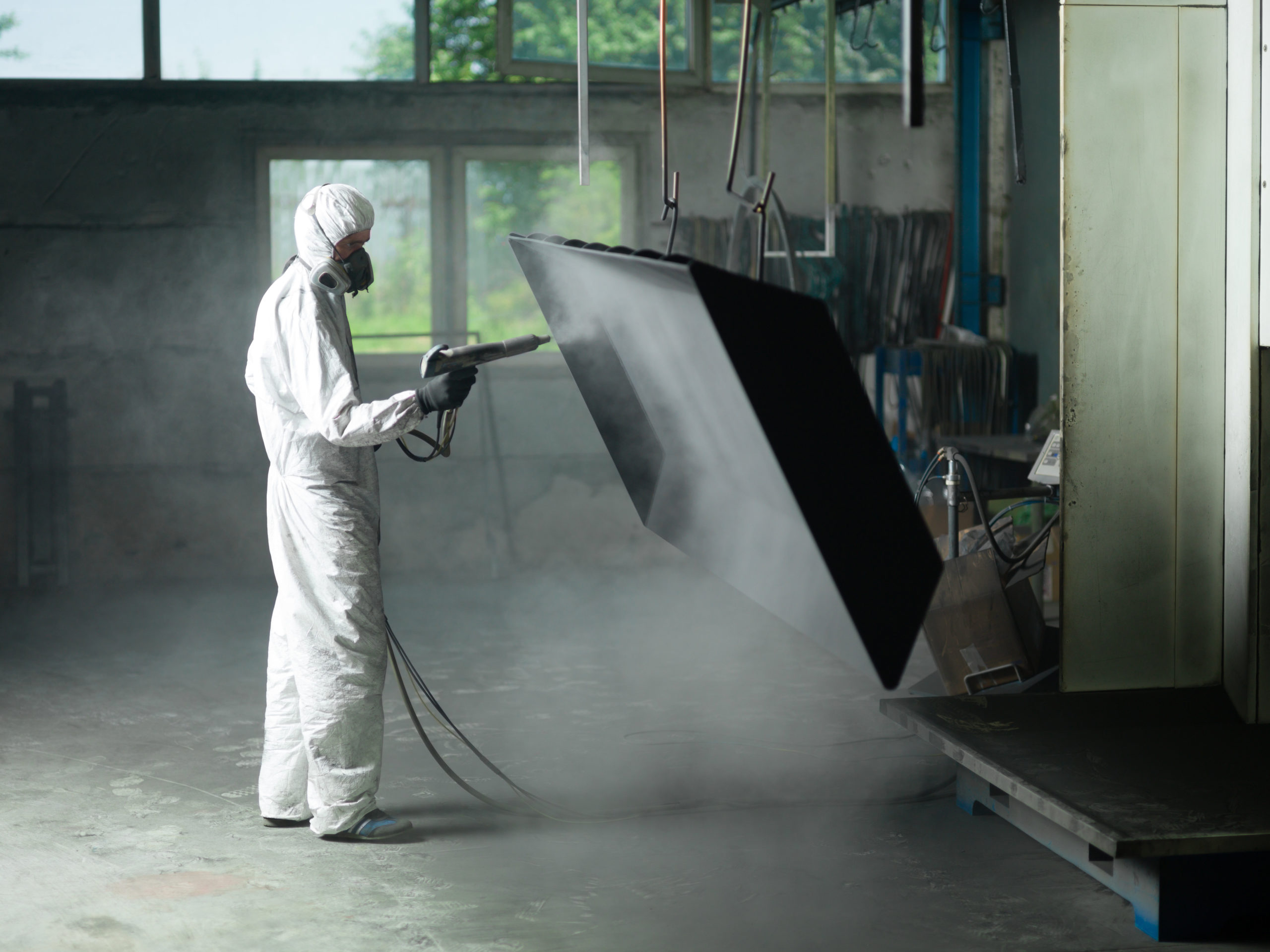 view of a worker wearing a full white protective suit and breathing mask, sand blasting a metal crate hung from a metal beam in the ceiling of an industrial hall